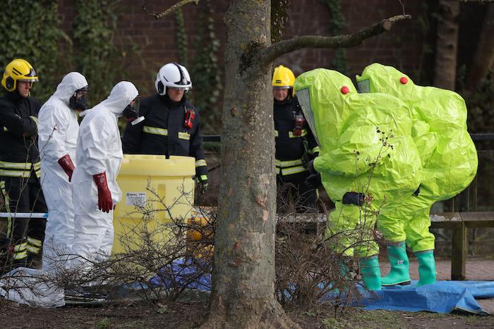 Firefighters in uniform and people in hi-viz hazmat suits stand in a semicircle looking at something on the floor that is obscured by a tree